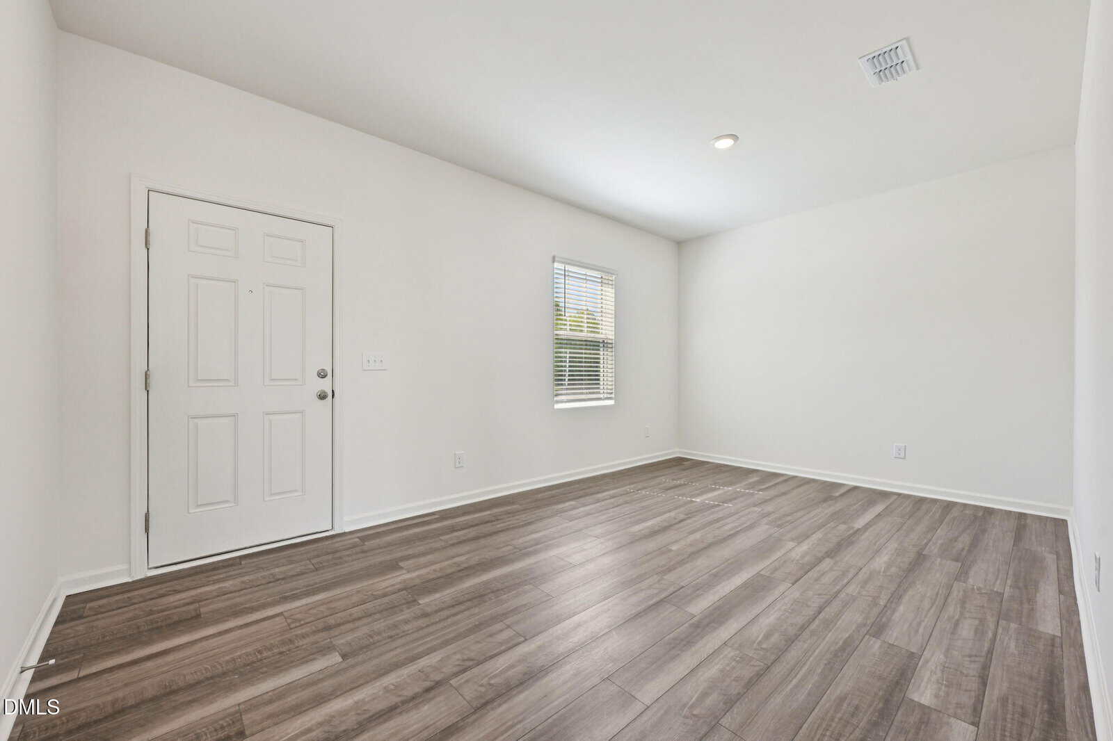 A bright, empty room with light wood flooring, white walls, a white front door, and a window letting in natural light.