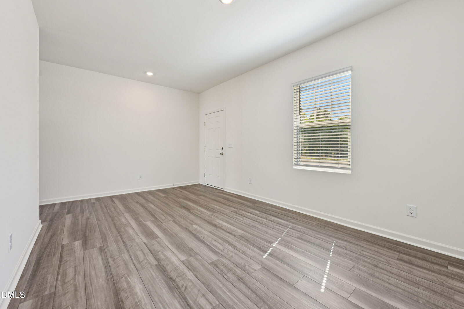 Bright, empty room with light-colored walls, wooden flooring, a window with blinds letting in sunlight, and a closed white door on the far wall. Ceiling lights are turned on.
