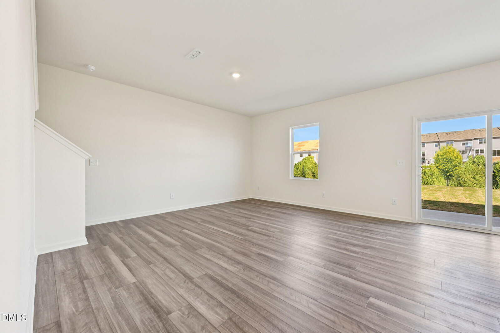 Bright, empty living room with light gray wood flooring, white walls, two windows, and a glass door leading to a grassy outdoor area; stairs partially visible on the left.