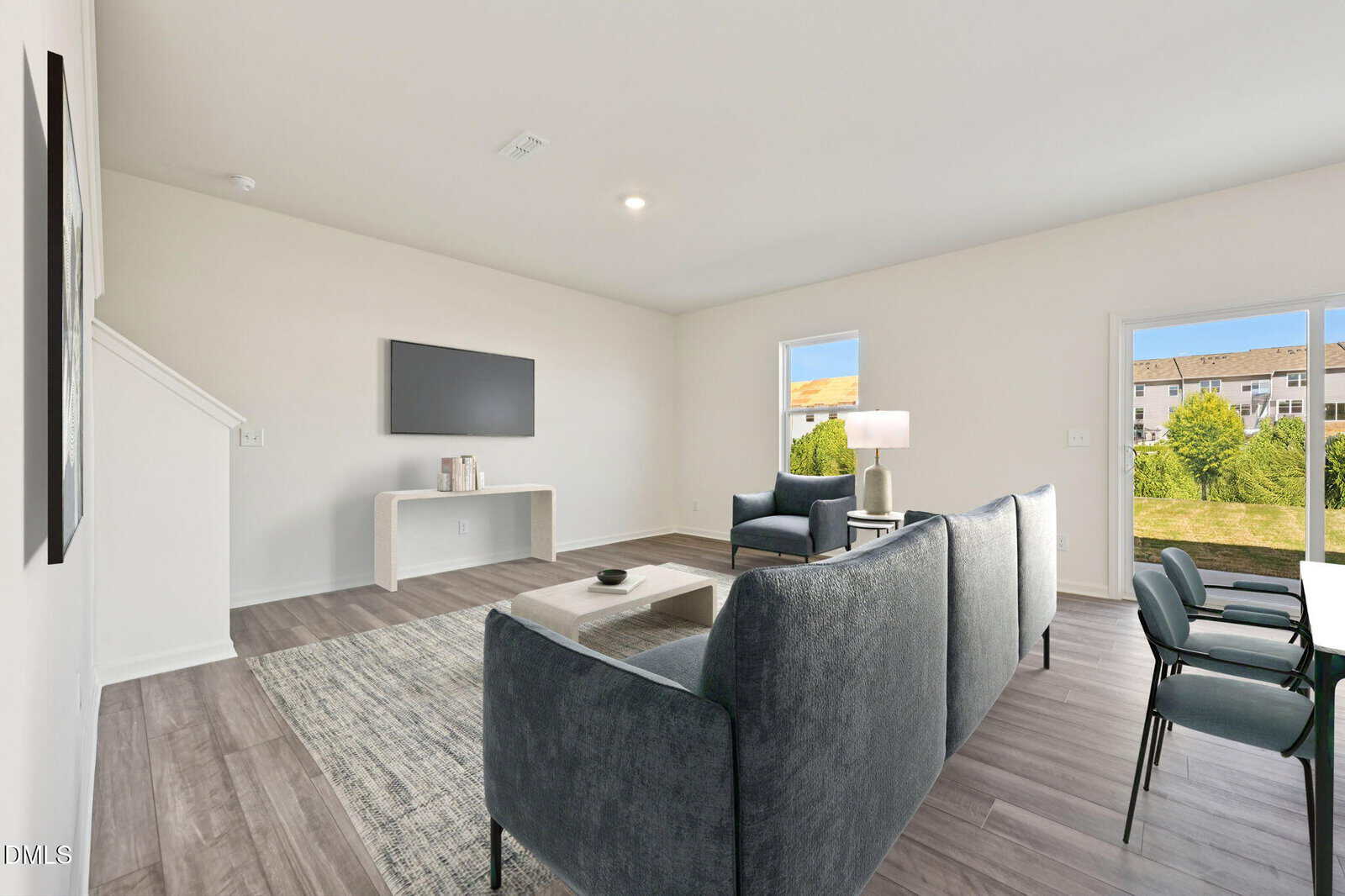 A modern living room with gray furniture, a wall-mounted TV, large windows showing greenery, and light wood flooring. The space is bright and minimalistic, with a neutral color palette and contemporary decor.