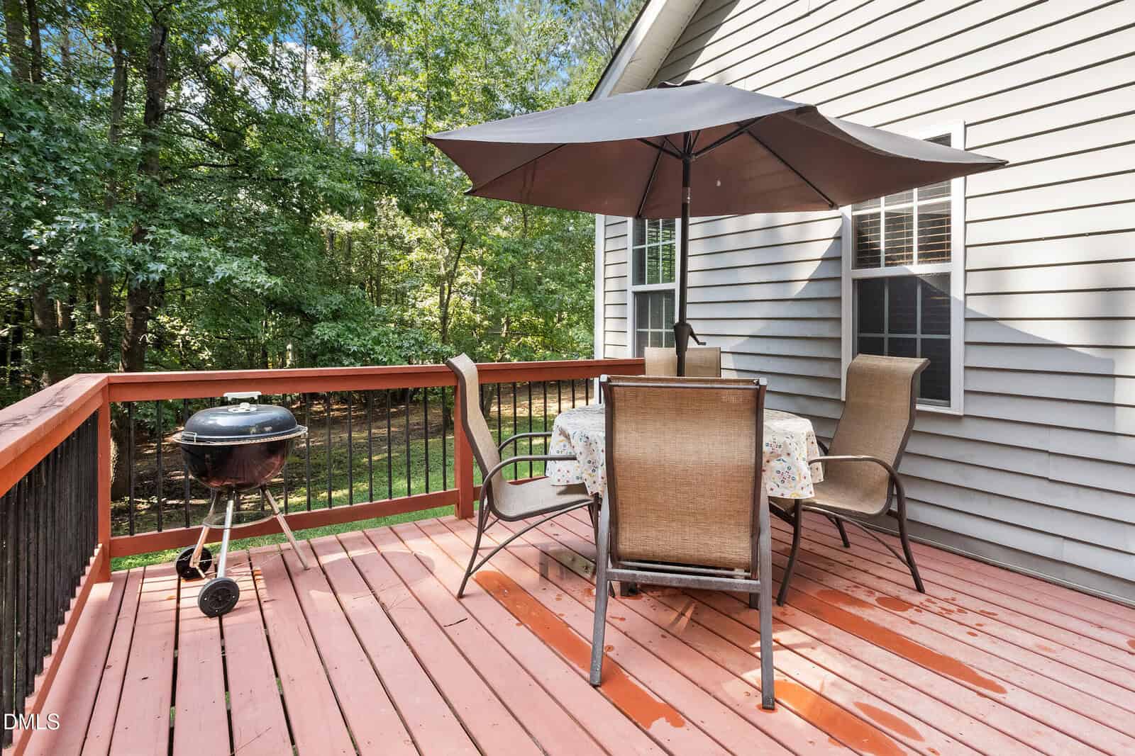 A wooden deck with a round grill, a table with a floral tablecloth, four chairs, and a large patio umbrella. The deck overlooks a wooded area and is attached to a light gray house.