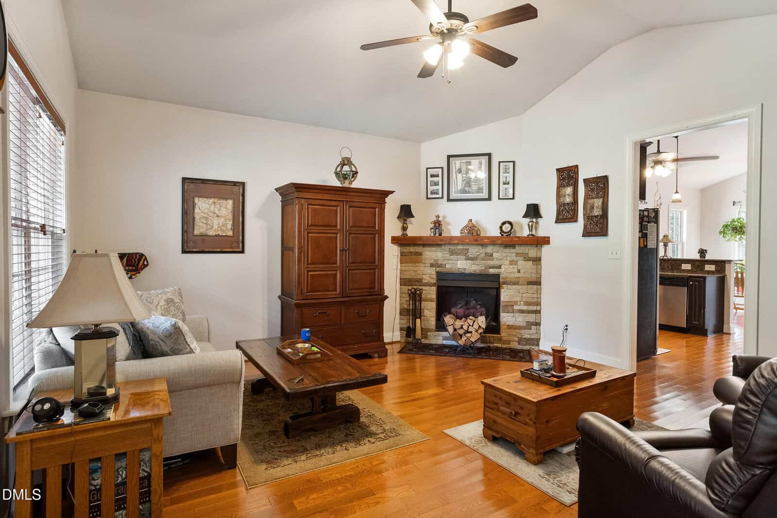 A cozy living room with hardwood floors, a beige sofa, wooden furniture, stone fireplace, and ceiling fan. Art and lamps decorate the space, with a view into the kitchen area.