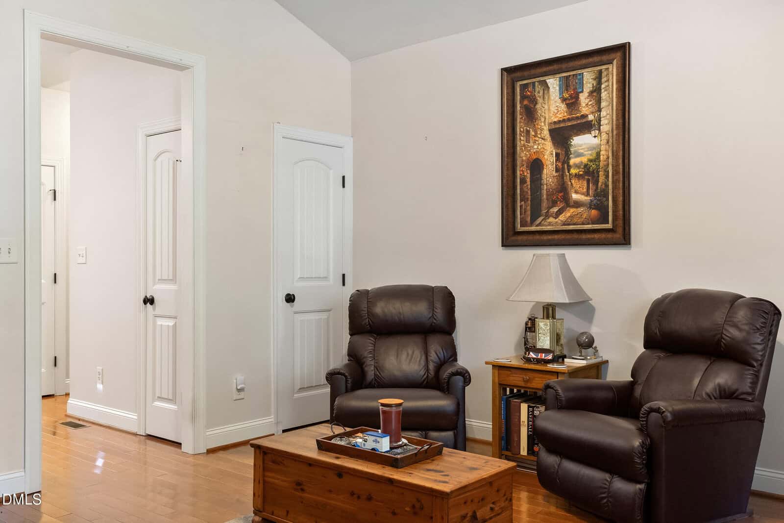 Two brown leather recliners face a wooden chest used as a coffee table, with a side table and lamp between them. A framed painting hangs on the wall above the arrangement.