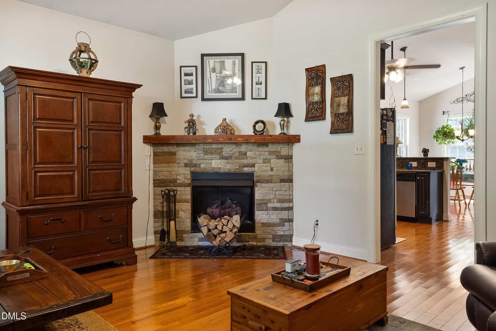 Living room with a stone fireplace, wooden mantel, and decor. A wooden armoire and chest sit on hardwood floors. Adjacent room has a ceiling fan and visible kitchen, with plants hanging by the window.