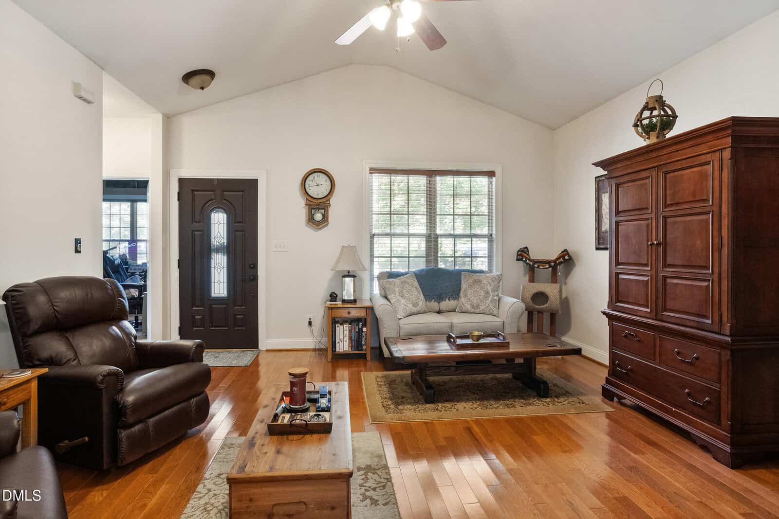 A cozy living room with wood floors, a dark armchair, sofa, coffee table, and a large wooden cabinet. A window lets in light, and a clock hangs on the wall above a small side table.