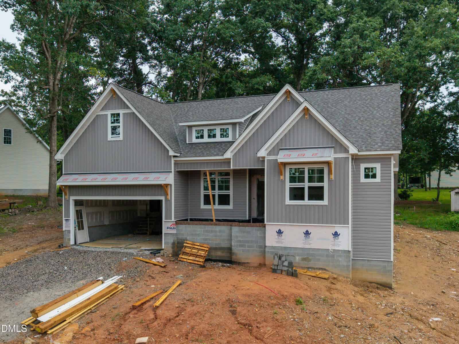 A gray two-story house under construction at 1039 Heather Lane, Wake Forest, with exposed foundation and materials scattered in the dirt yard. The garage door is open, and some siding and windows are not yet finished. Trees are visible in the background.