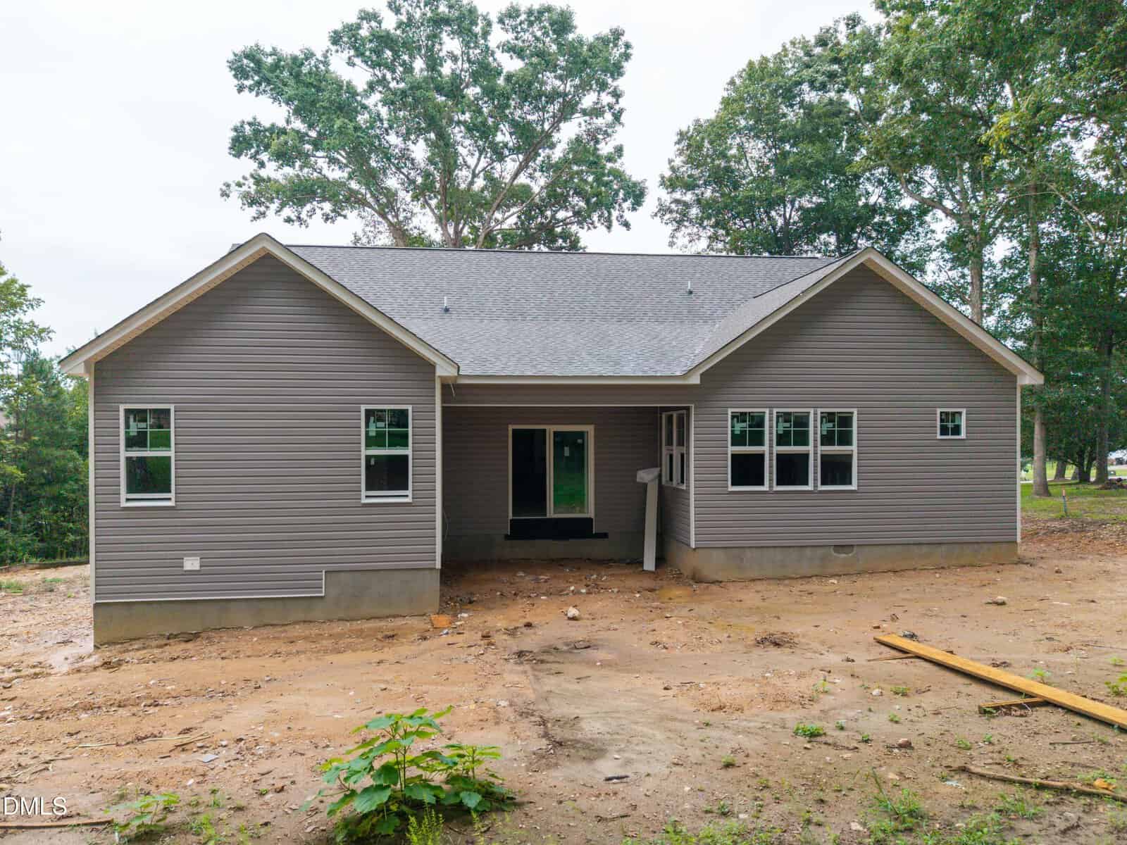 A single-story gray house with white trim and multiple windows is pictured on a dirt lot. The yard is unfinished, and trees are visible in the background. The house has a covered back patio area.