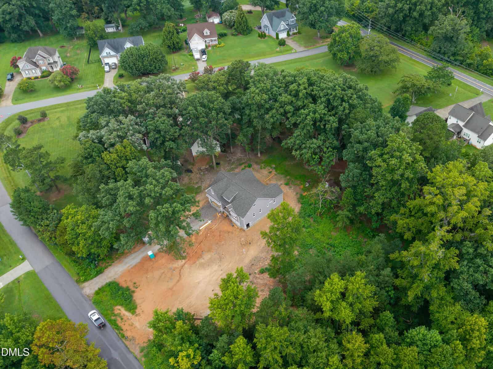 Aerial view of a house under construction surrounded by trees, with dirt clearing around the building. Nearby paved roads and several completed houses are visible in a wooded suburban neighborhood.