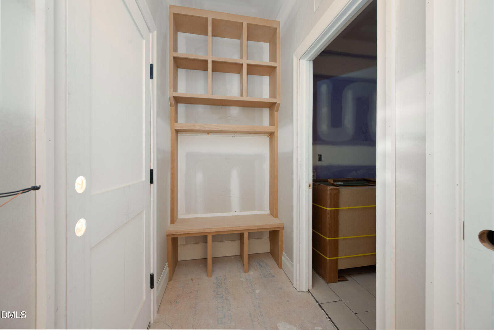 A hallway with white walls features built-in wooden shelves and a bench. There are open cubby spaces above and below the bench. Two adjacent doorways are visible on the right and left sides.
