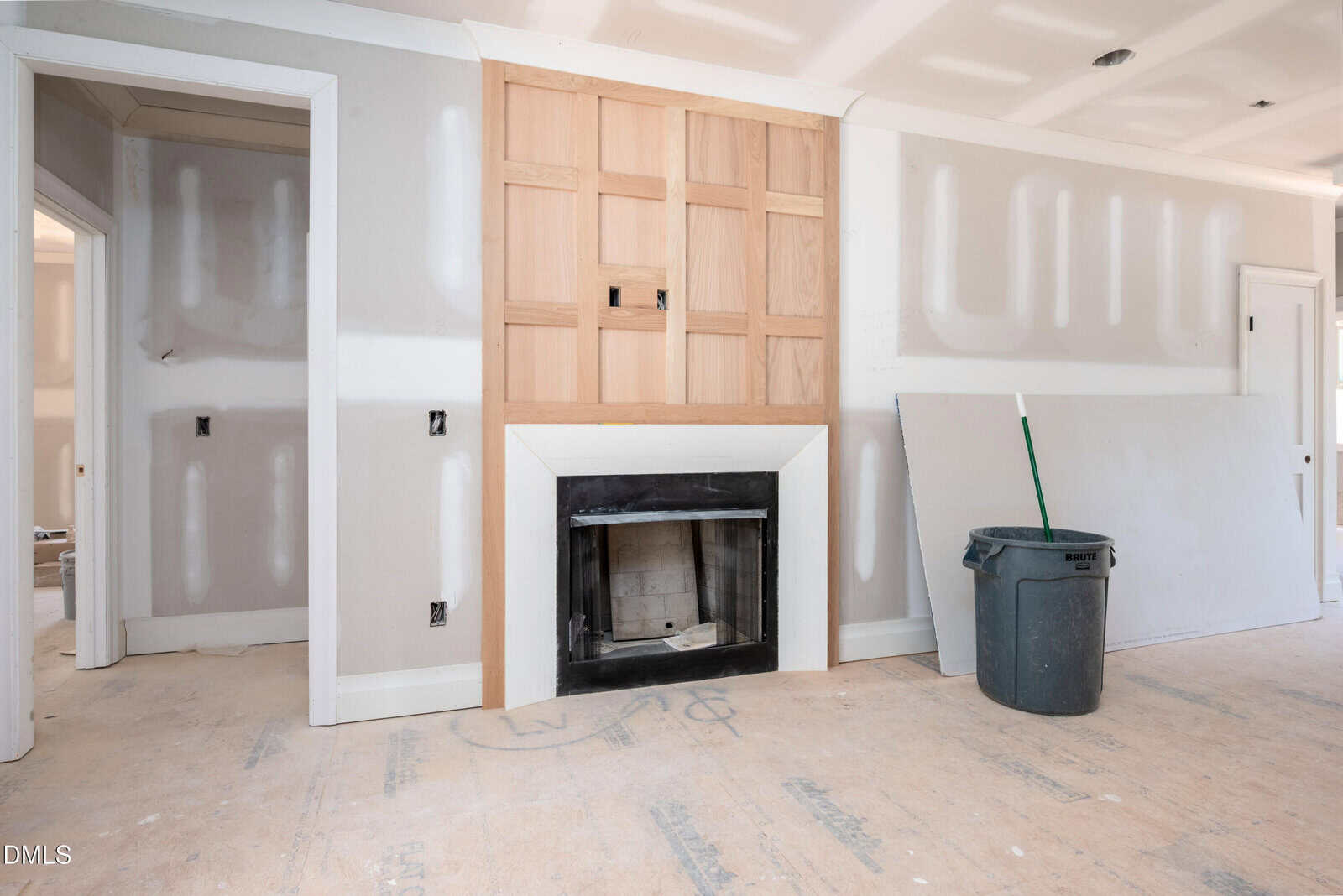 A room under construction with unfinished walls, a fireplace with wood paneling above it, and a trash can with a broom nearby. The floor is covered with protective material.