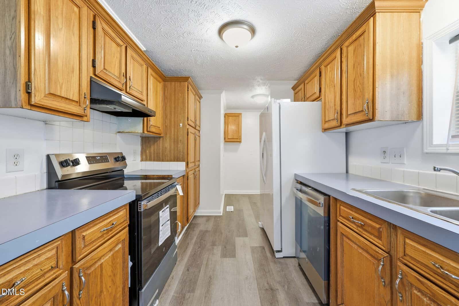 Narrow kitchen with wood cabinets, light gray countertops, stainless steel appliances, white tile backsplash, and light wood flooring. Ceiling lights brighten the space. A window is above the sink on the right.