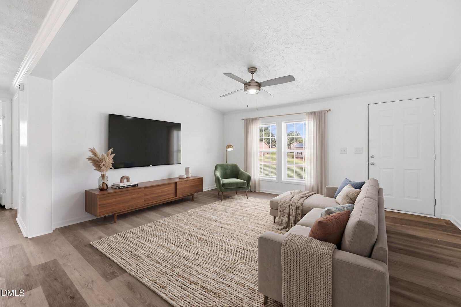 Modern living room with light walls, a large wall-mounted TV, a wooden console, a green armchair, a beige sofa, a textured rug, and large windows letting in natural light.