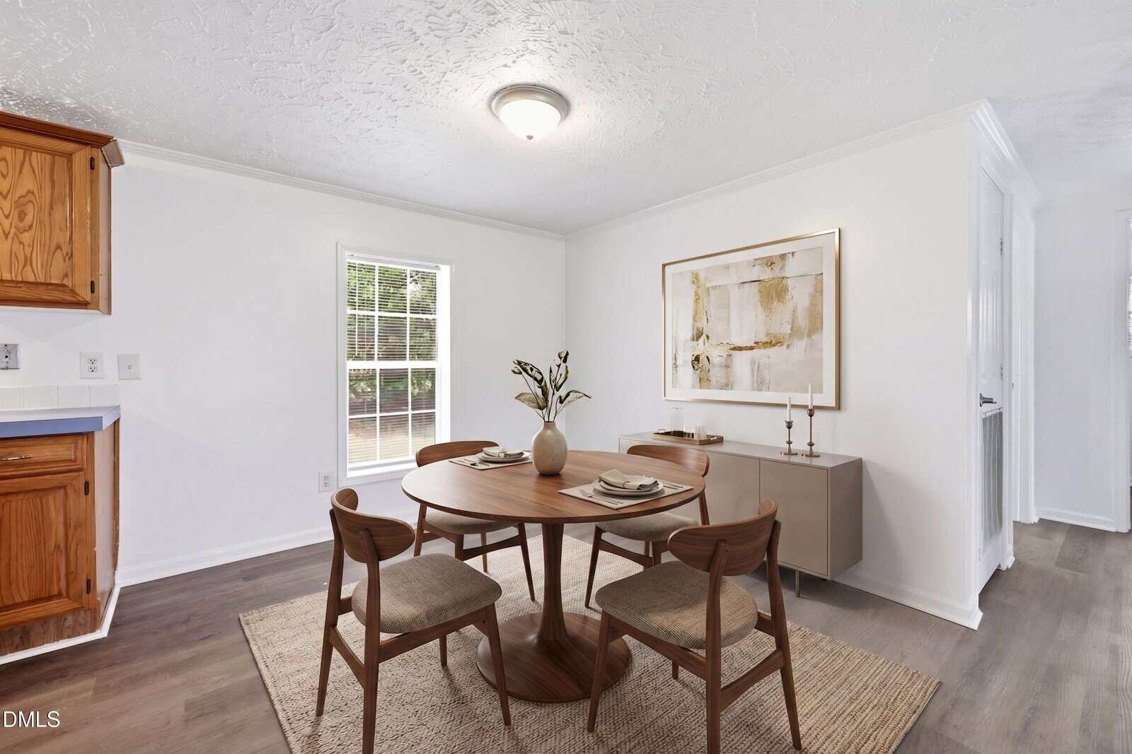 A small dining area with a round wooden table, four chairs, a vase centerpiece, and a sideboard under a large abstract painting. Natural light comes through a window, highlighting the neutral decor and wooden accents.