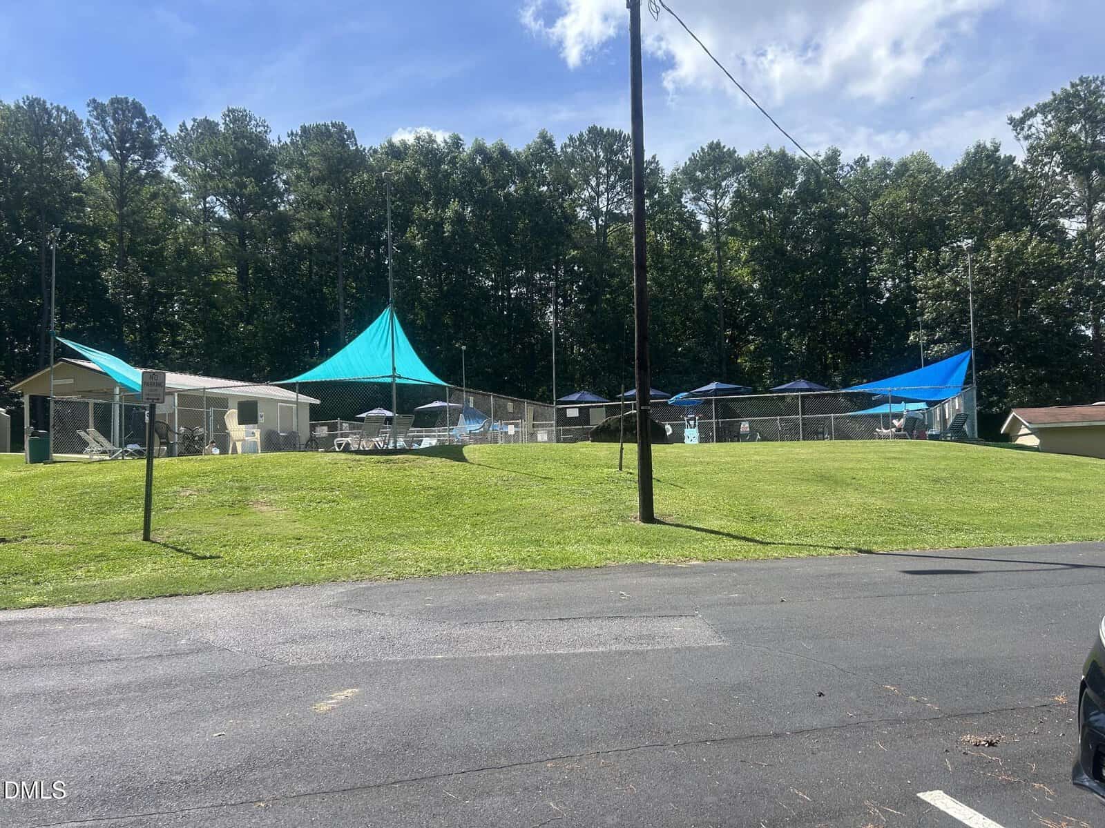 A fenced outdoor pool area with blue sun shades, lounge chairs, umbrellas, and a small building, surrounded by grass, trees, and a paved parking area under a partly cloudy sky.