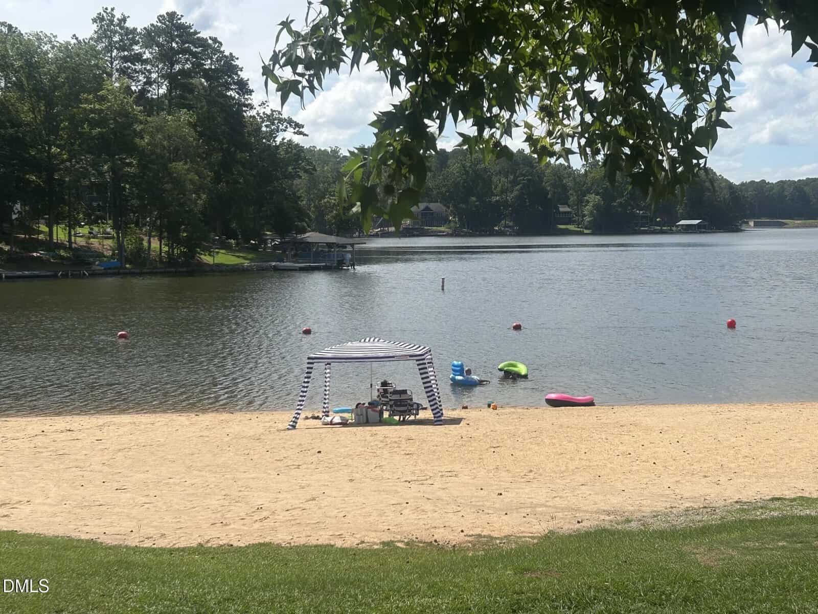 A sandy beach by a lake with a striped canopy, chairs, and colorful floats. Trees and houses line the lake shore under a partly cloudy sky.