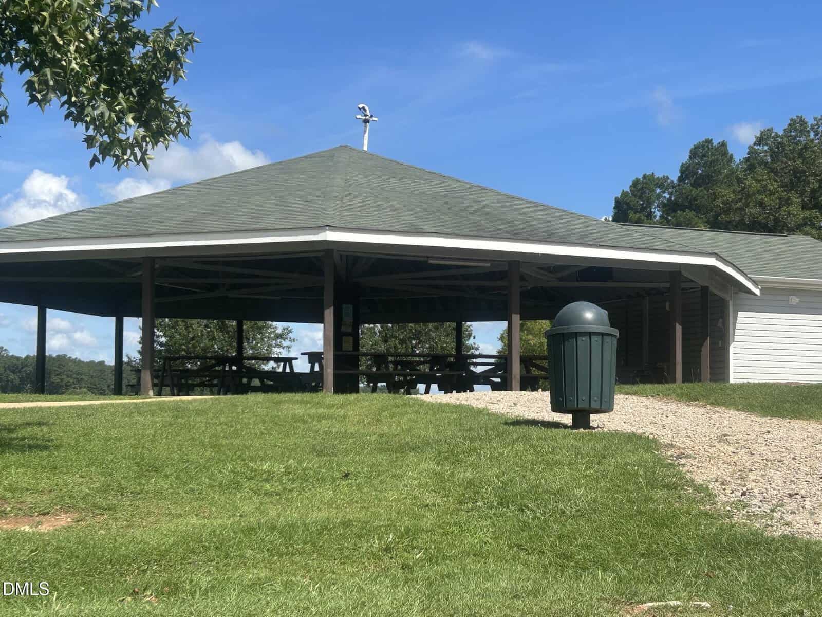 A large open-air pavilion with picnic tables sits on green grass under a blue sky. A trash can and gravel path are in the foreground, and trees are visible in the background.