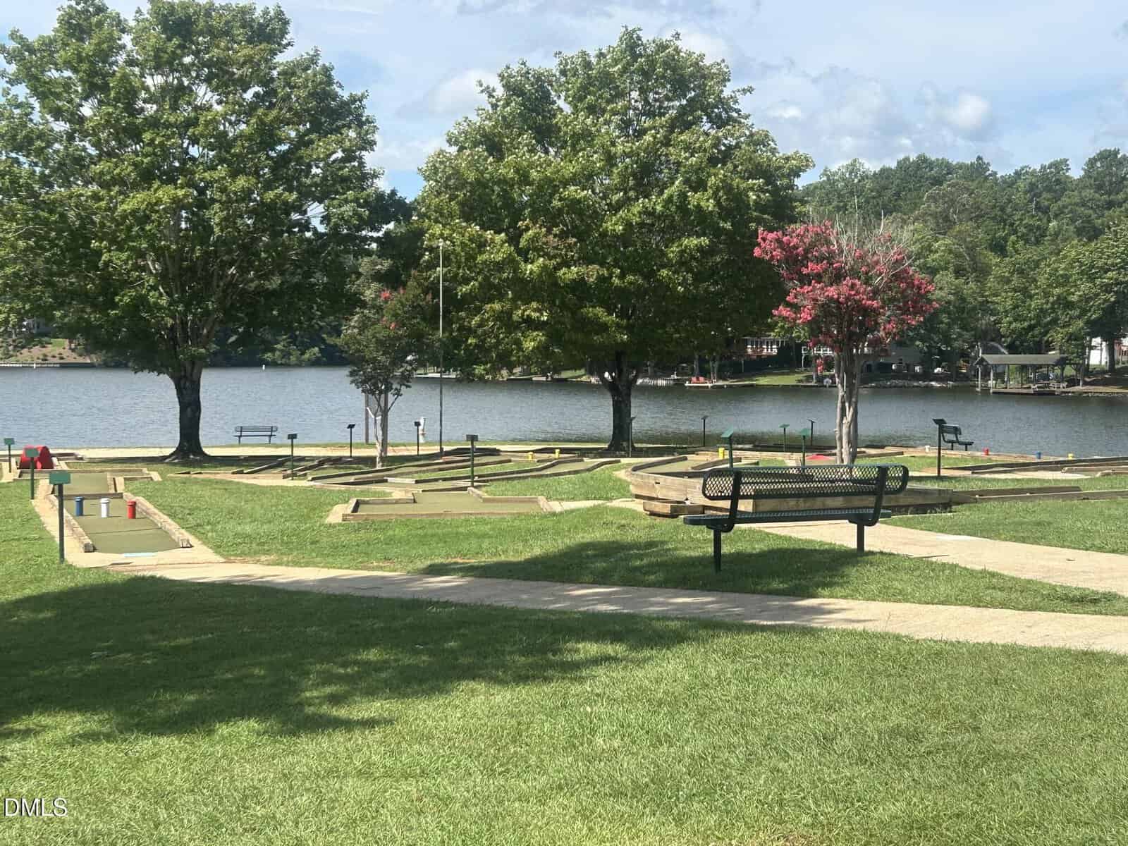 A mini-golf course with green lawns and trees sits beside a calm lake. Benches and walking paths are visible, with houses and more trees in the background under a partly cloudy sky.
