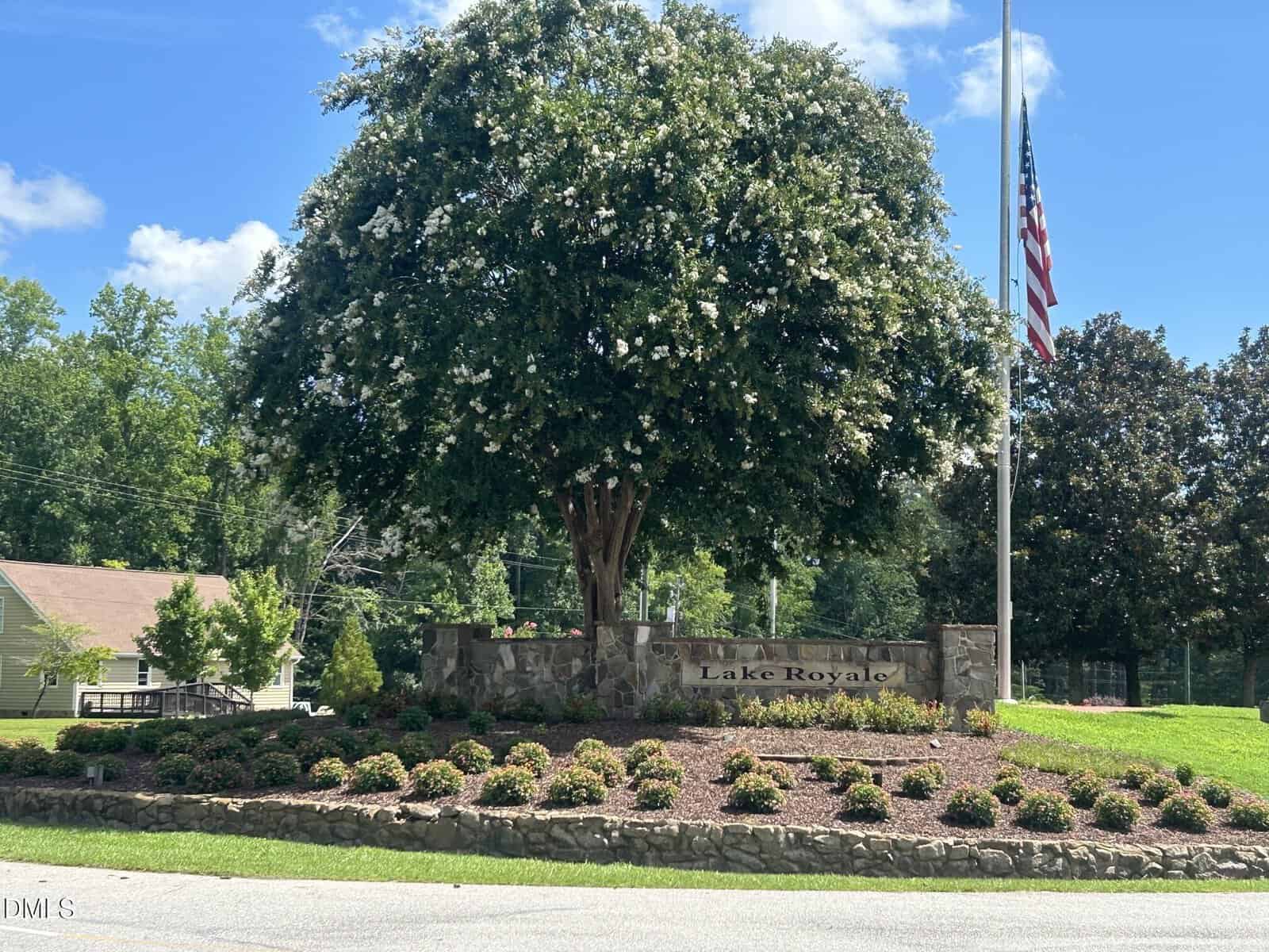 A large tree with white flowers stands behind a stone sign reading "Lake Royale" in Louisburg, surrounded by landscaped bushes and a nearby American flag on a sunny day near 116 Buckaroo Drive.