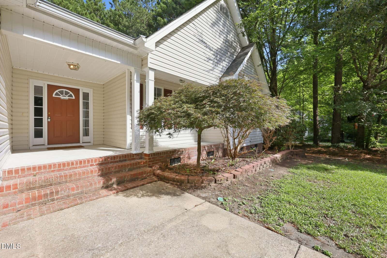 A house with beige siding, a red front door, brick steps, a covered porch, and a landscaped yard with a small tree and shrubs, surrounded by tall green trees.