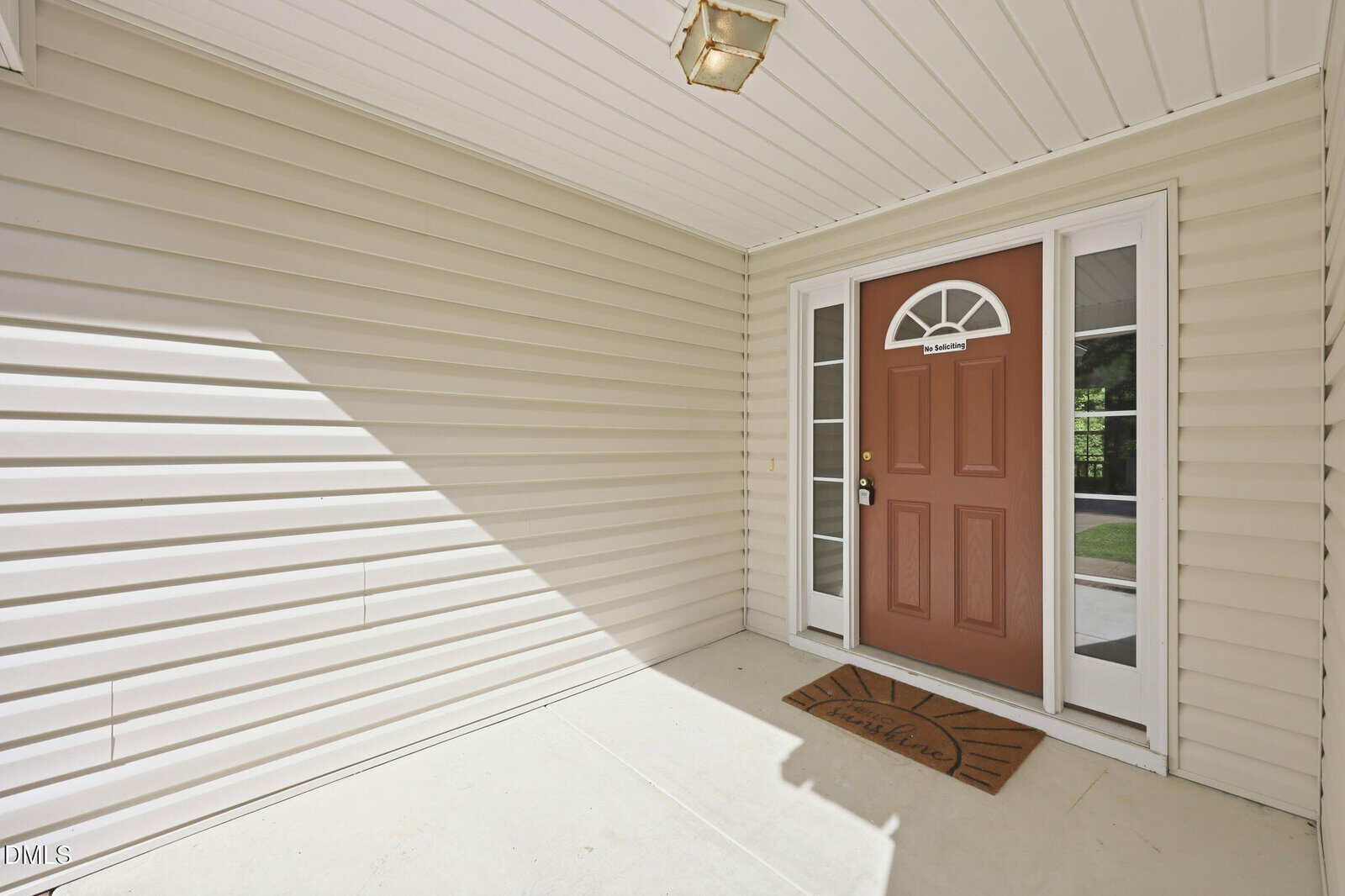 Covered front porch with cream-colored siding, a brown front door featuring a half-window, two narrow side windows, a ceiling light, and a welcome mat placed in front of the entrance.