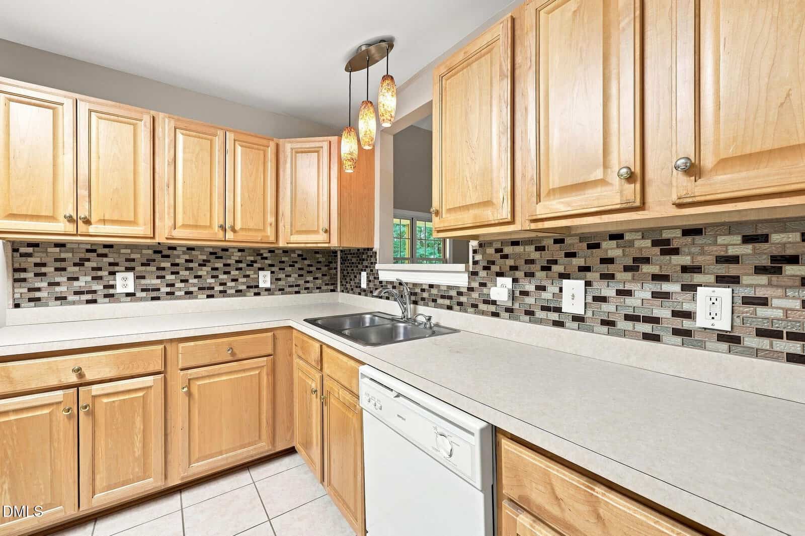 Modern kitchen with light wood cabinets, white countertops, mosaic tile backsplash, dishwasher, stainless steel sink, pendant lights, and a window above the sink.