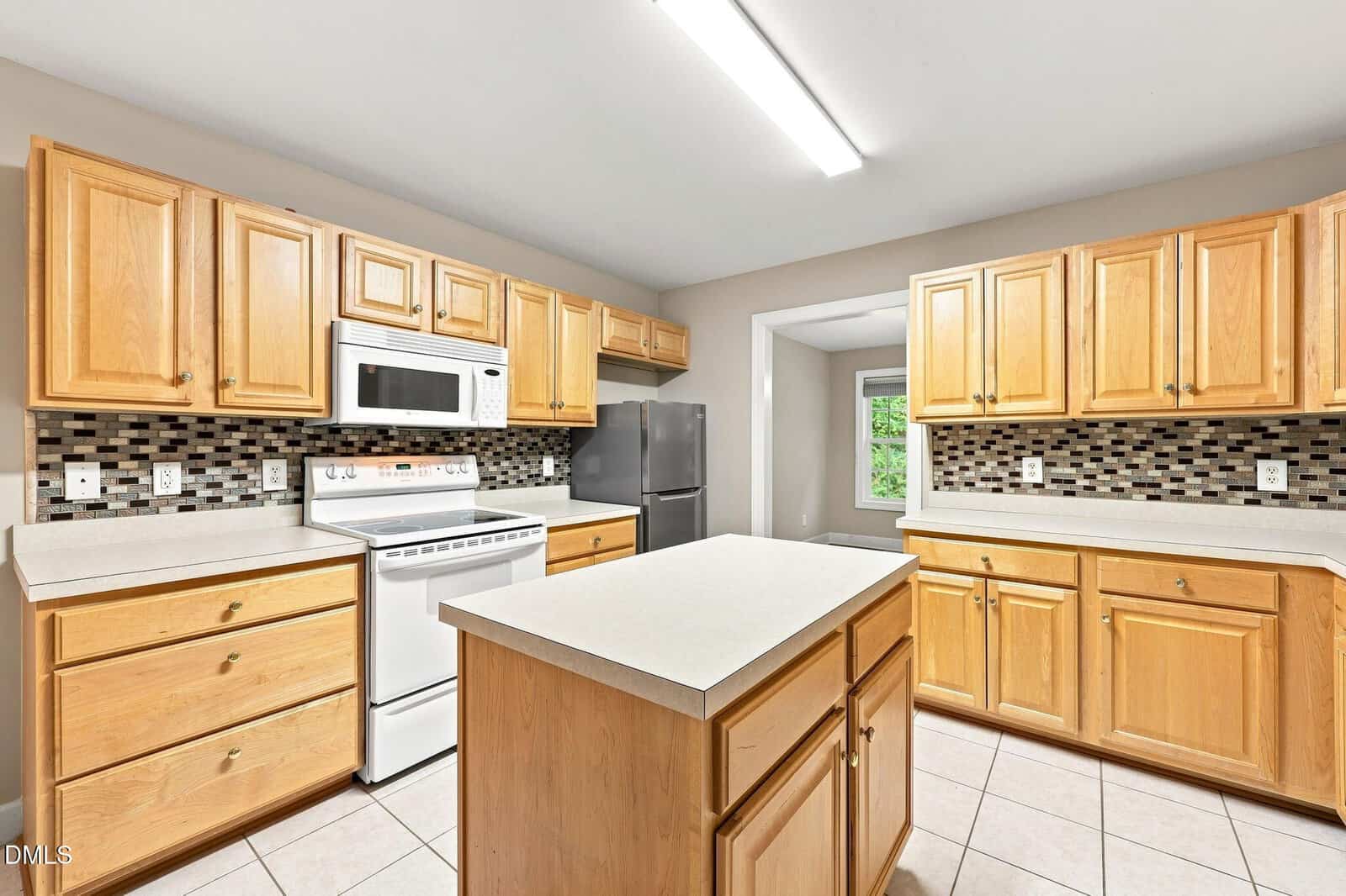 Modern kitchen with light wood cabinets, white countertops, a central island, stainless steel fridge, white stove and microwave, and a mosaic tile backsplash under bright overhead lighting.