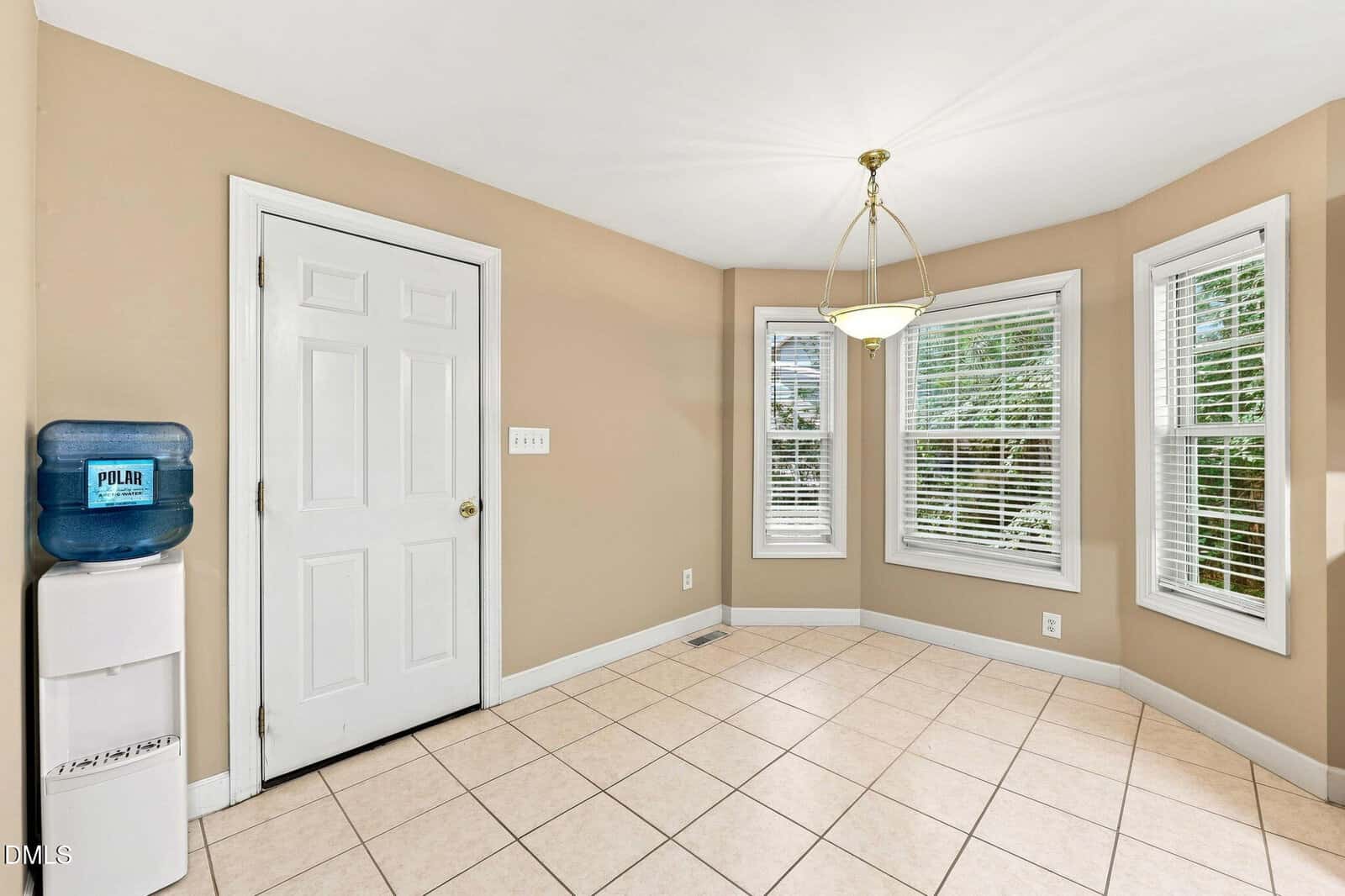 A bright room with beige walls, white tile floor, a white door, a small water dispenser, a brass light fixture, and three large bay windows with white blinds.