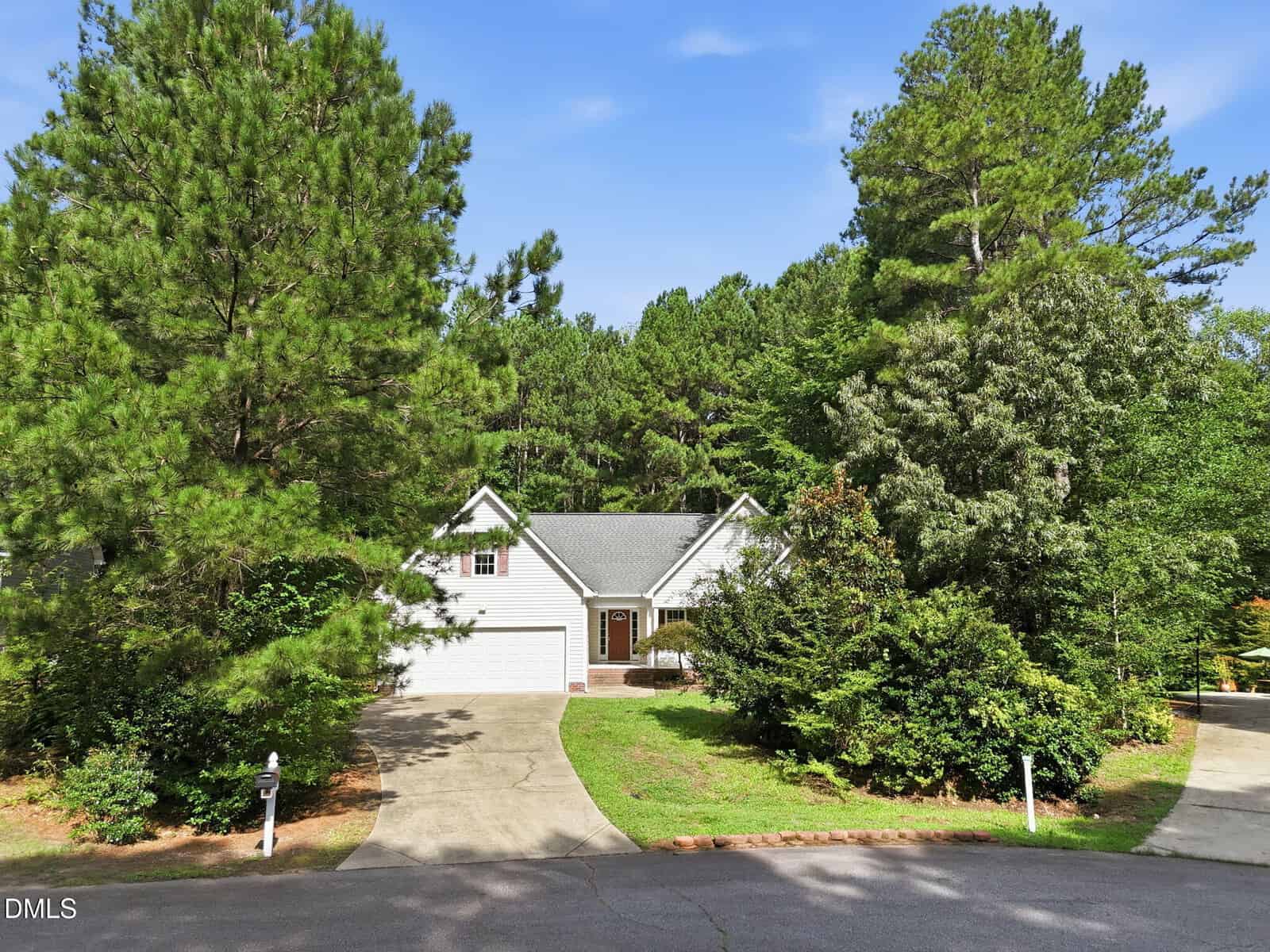 A white house with a gray roof, two-car garage, and driveway sits surrounded by tall green trees and bushes on a quiet, curved street under a clear blue sky.