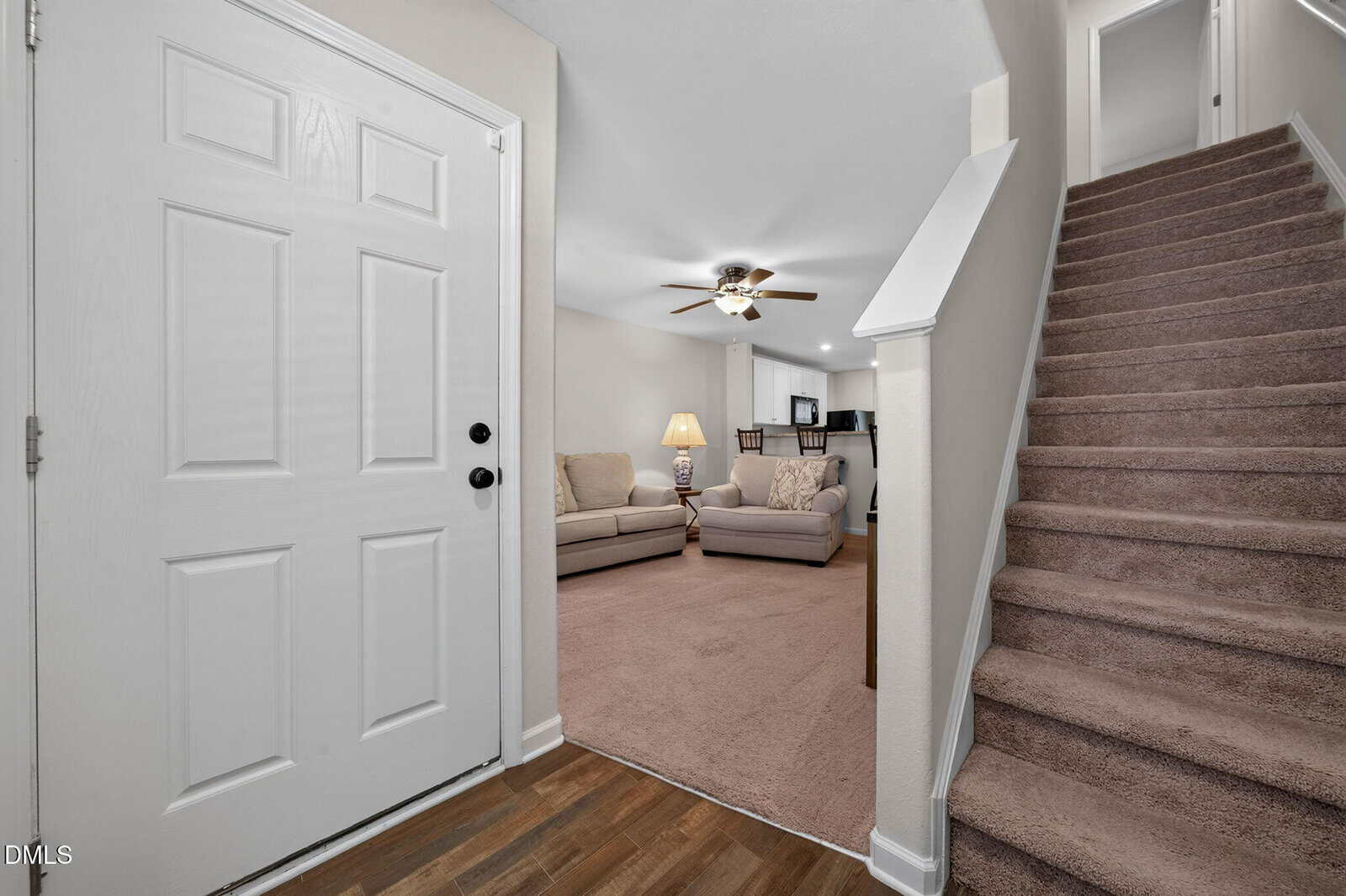 View of a home entryway with a white front door, brown carpeted stairs, and a living room featuring beige sofas, a lamp, ceiling fan, and a TV in the background. Walls are light-colored with wood and carpet flooring.