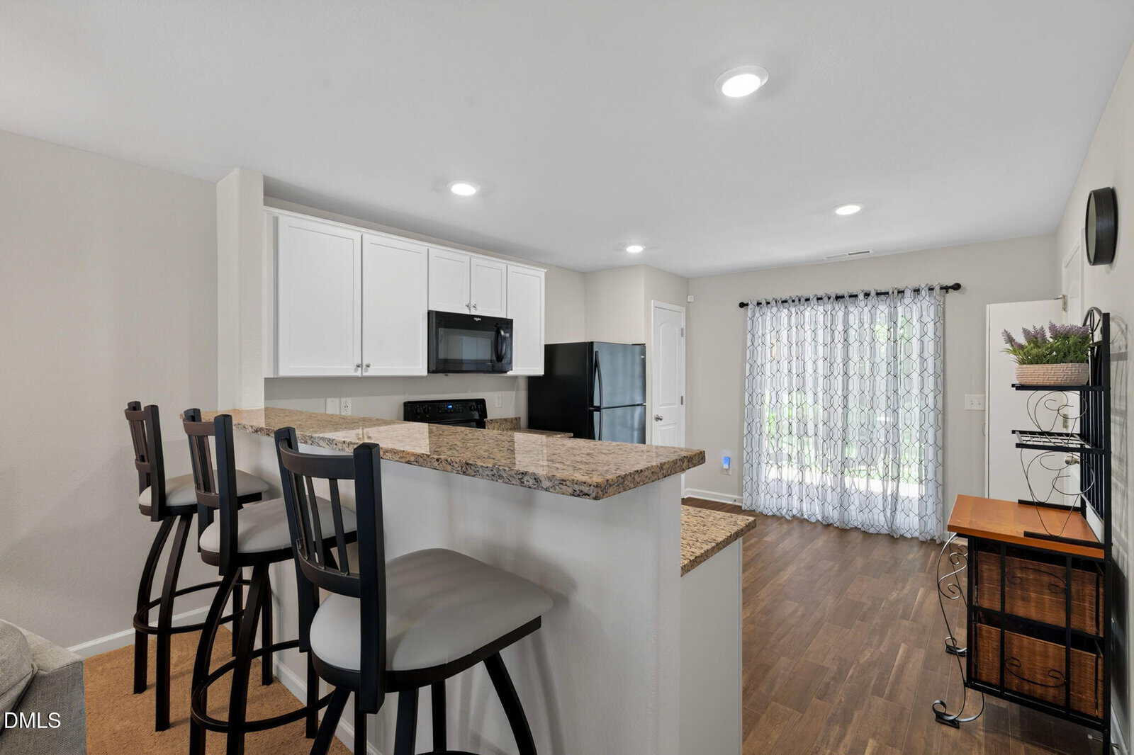 Modern kitchen with granite countertops, three bar stools, white cabinets, black appliances, and wood flooring. Sunlight enters through glass doors with sheer curtains, and a small table with a plant sits near the window.