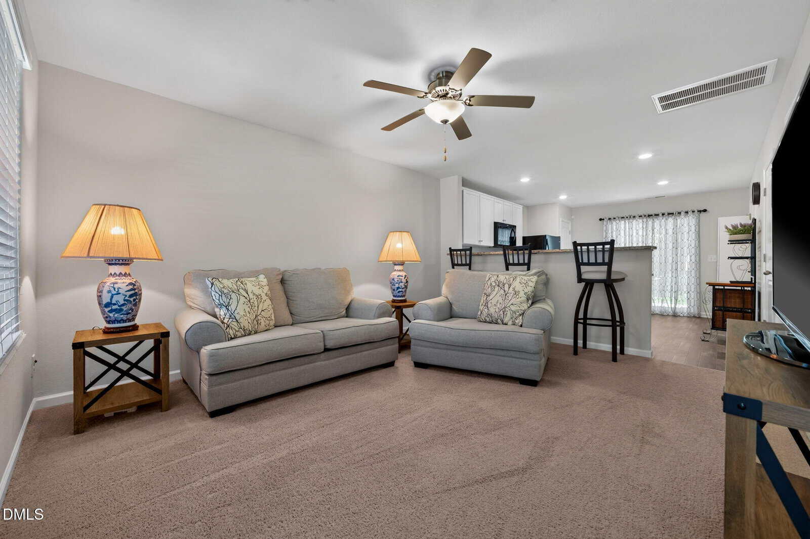 A cozy living room with two gray sofas, decorative pillows, two blue-patterned lamps, a ceiling fan, and a breakfast bar with three stools. Carpeted floor and neutral walls create a warm, inviting atmosphere.