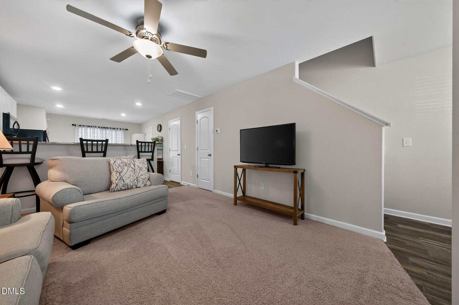 A cozy living room with beige carpet, a light gray sofa, a TV on a wooden stand, bar stools at a kitchen counter, and a ceiling fan overhead. An open staircase and white doors are visible on the right wall.