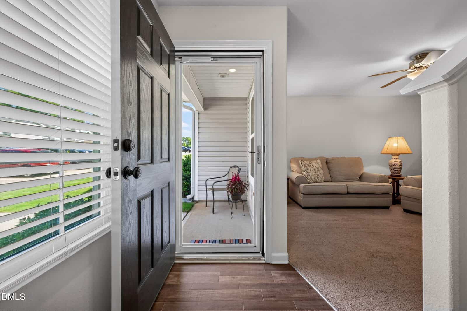 Open front door view of a home, leading to a covered porch with a chair and potted plant. Inside, a living room with beige couches, a lamp, and ceiling fan is visible, with carpeted and wood flooring.