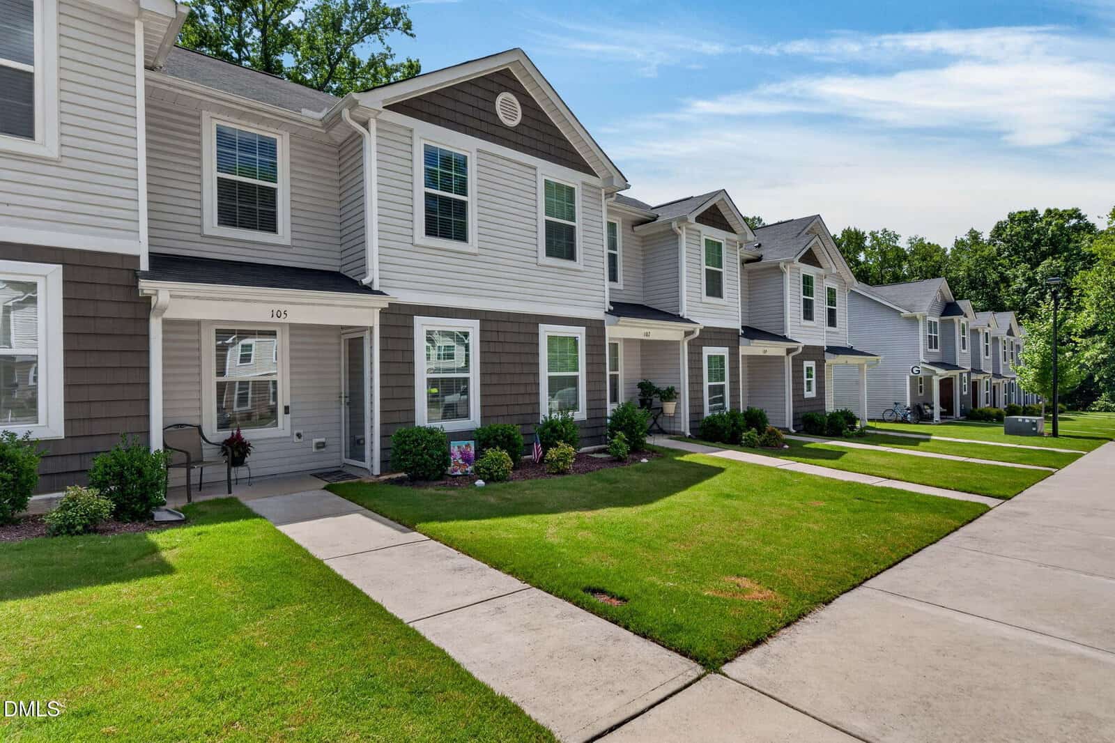 Row of modern two-story townhomes with gray siding, white trim, and small front lawns. Bright, sunny day with green grass and trees lining the sidewalk.