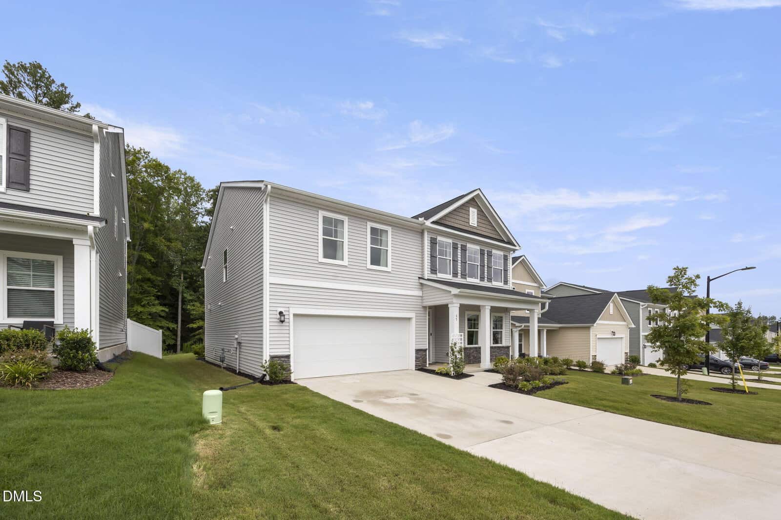 Two-story suburban house with gray siding, white trim, a two-car garage, and a wide driveway. Well-maintained green lawn and landscaping, neighboring houses, and a clear blue sky.