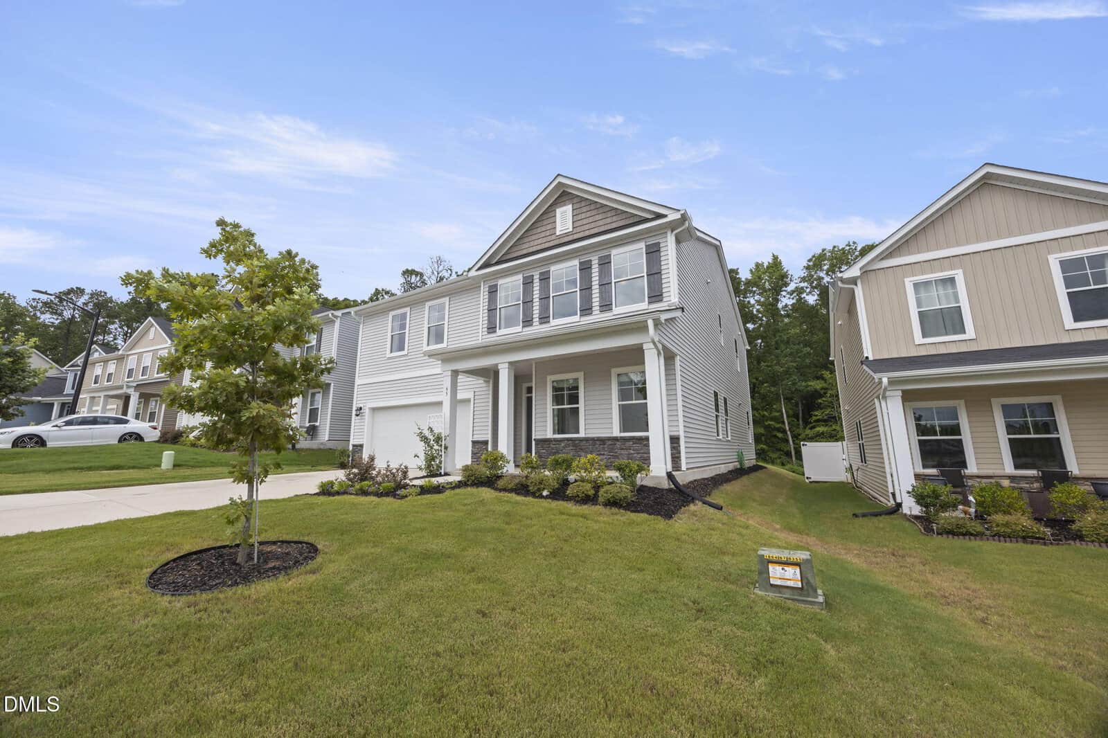 Two-story suburban house with light-colored siding, front porch, and attached garage sits on a sloped lawn with young trees and shrubs. A real estate sign is visible near the driveway, neighboring homes line the street.