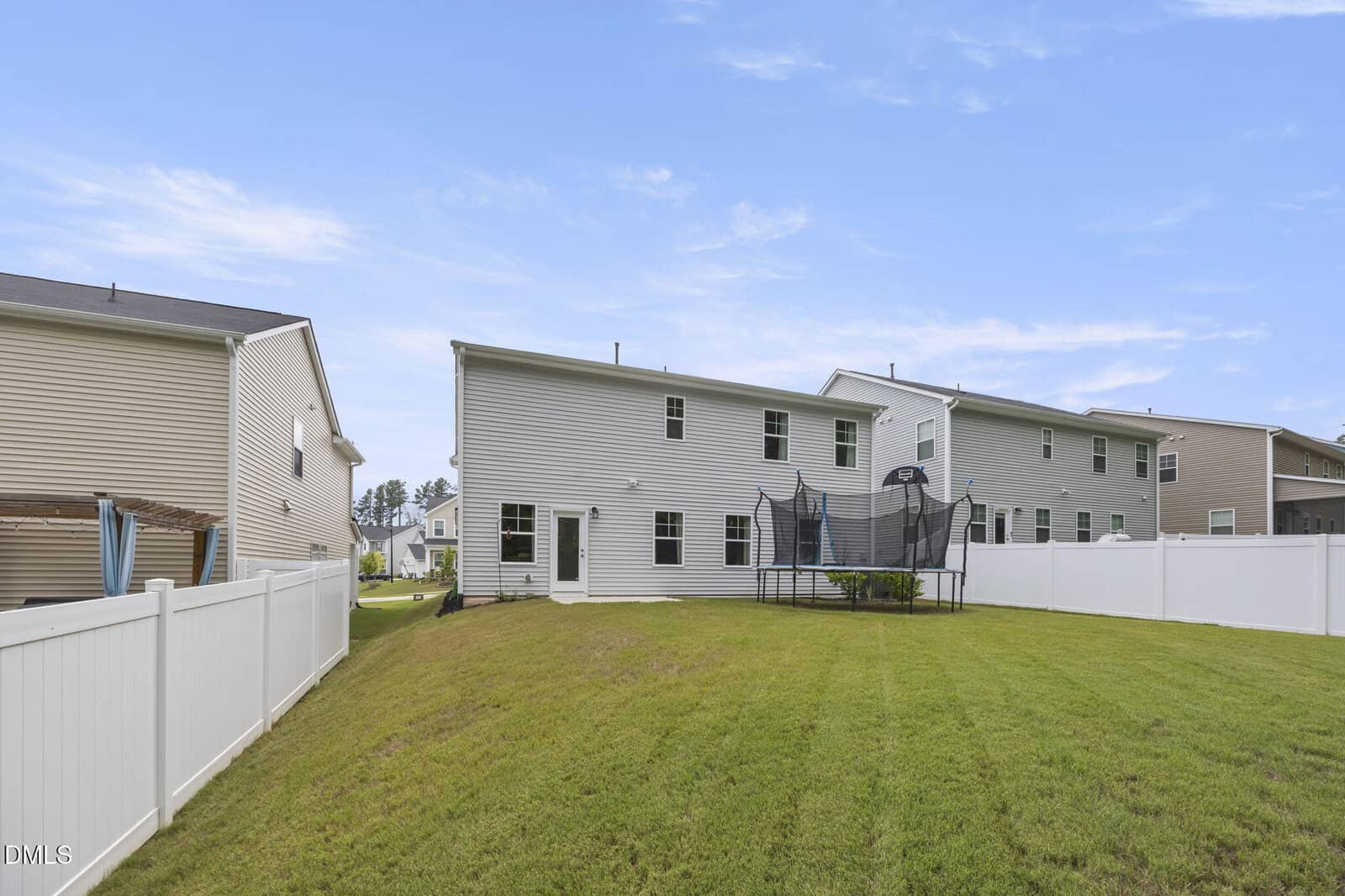 A fenced backyard with green grass, a white two-story house, neighboring homes, and a trampoline near the center of the yard under a blue, partly cloudy sky.