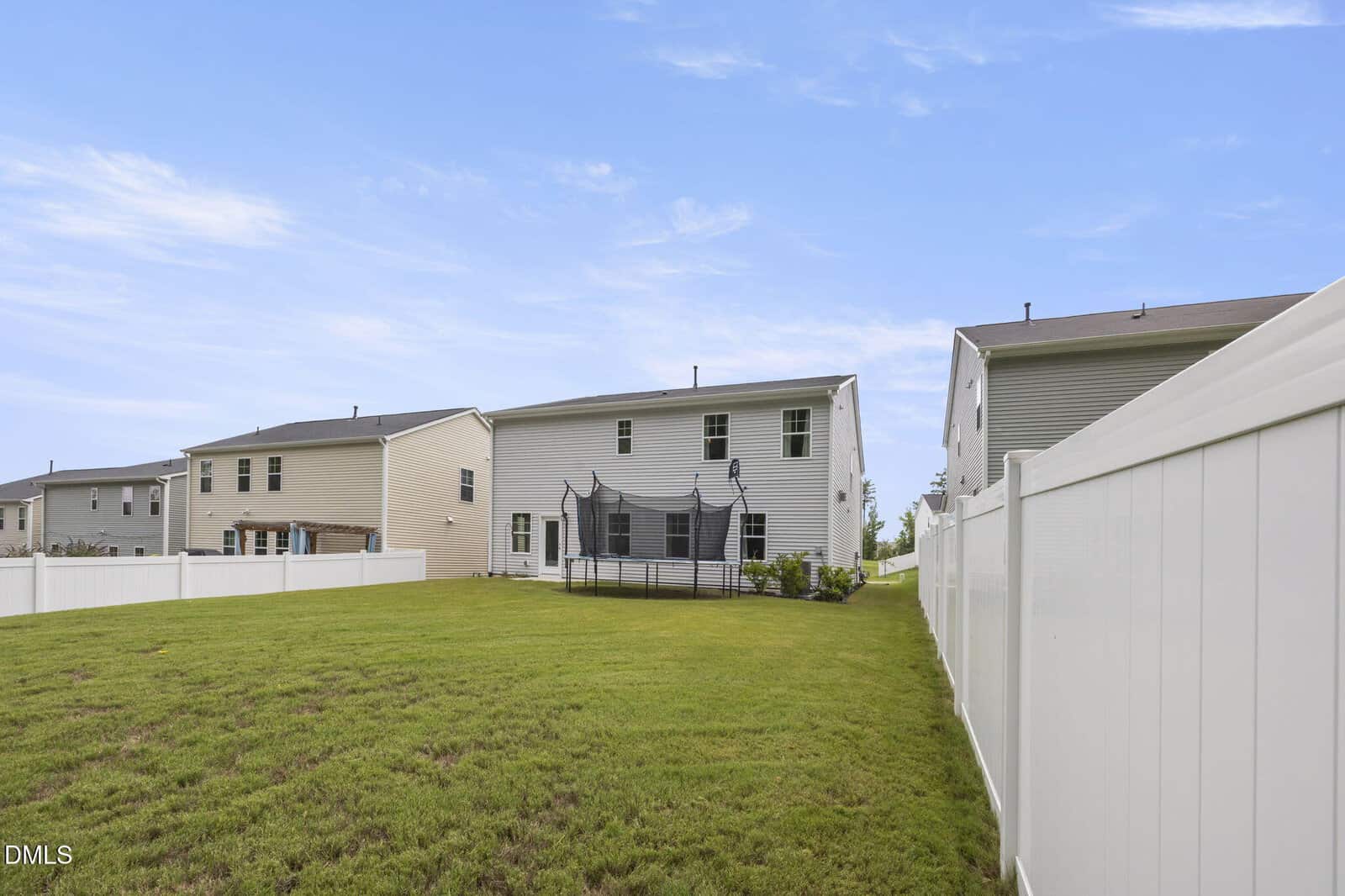 A fenced backyard with green grass, a trampoline, and two-story houses in the background under a blue sky with light clouds.