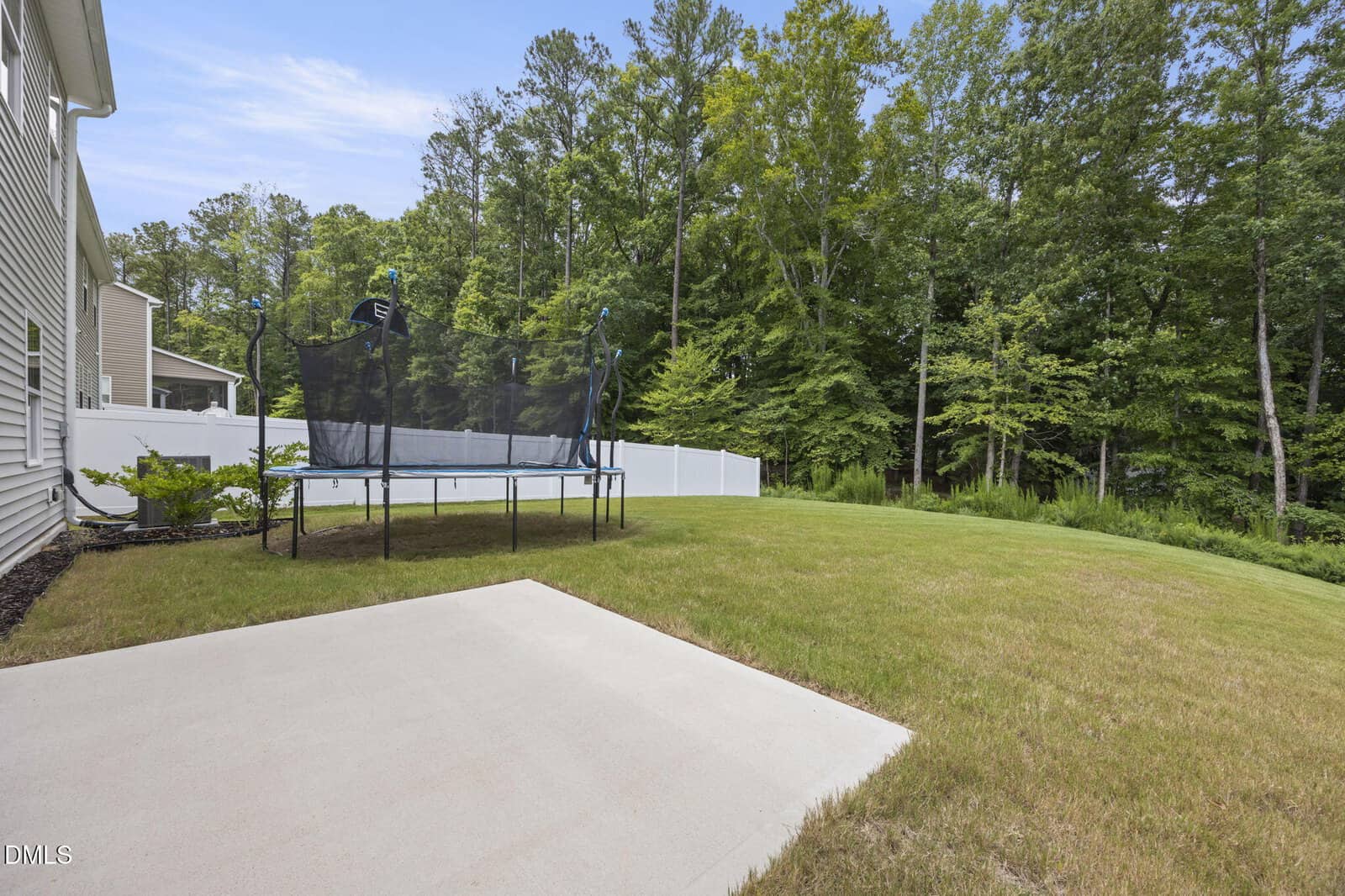 A backyard with a concrete patio, grassy lawn, black trampoline, white privacy fence, and tall trees in the background under a blue sky.