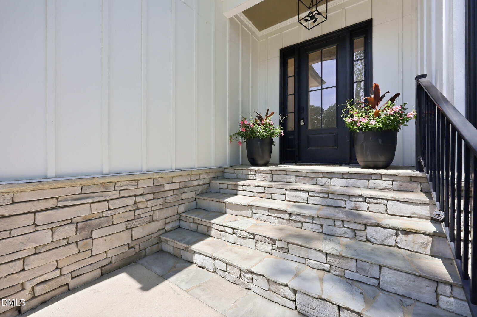 Stone steps lead to a black double door with glass panes. Two large potted plants flank the entrance. The house has white siding and a stone wall. A black lantern hangs above the door.