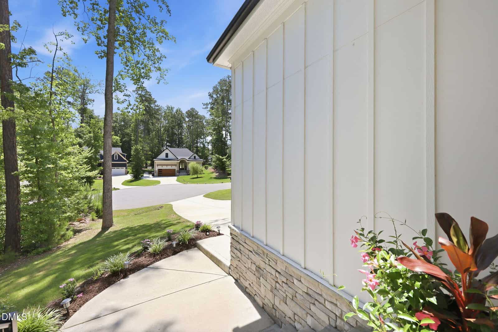 View from a front porch with white siding and stone accents, showing a curved sidewalk, landscaped yard, tall trees, and houses across a quiet suburban street under a clear blue sky.