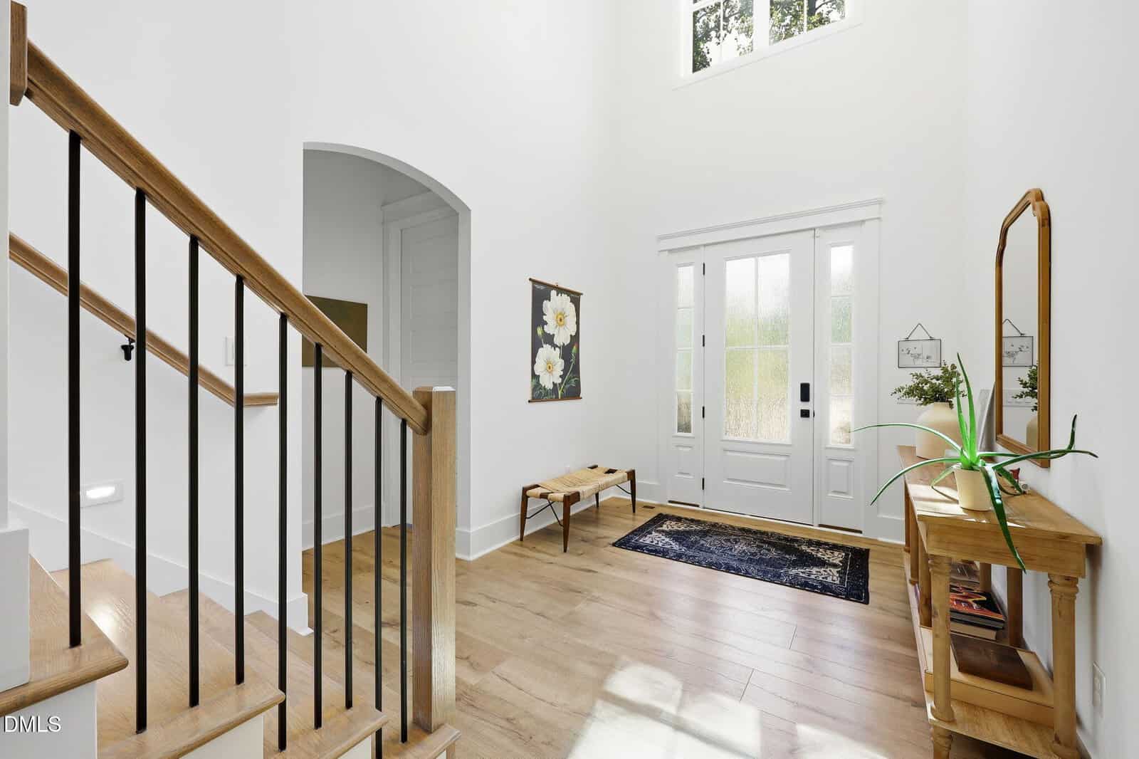 Bright entryway with light wood floors, white walls, a wooden staircase with black rails, a wooden console table with plants, a bench, floral artwork, and a black rug by the front door with glass panels.