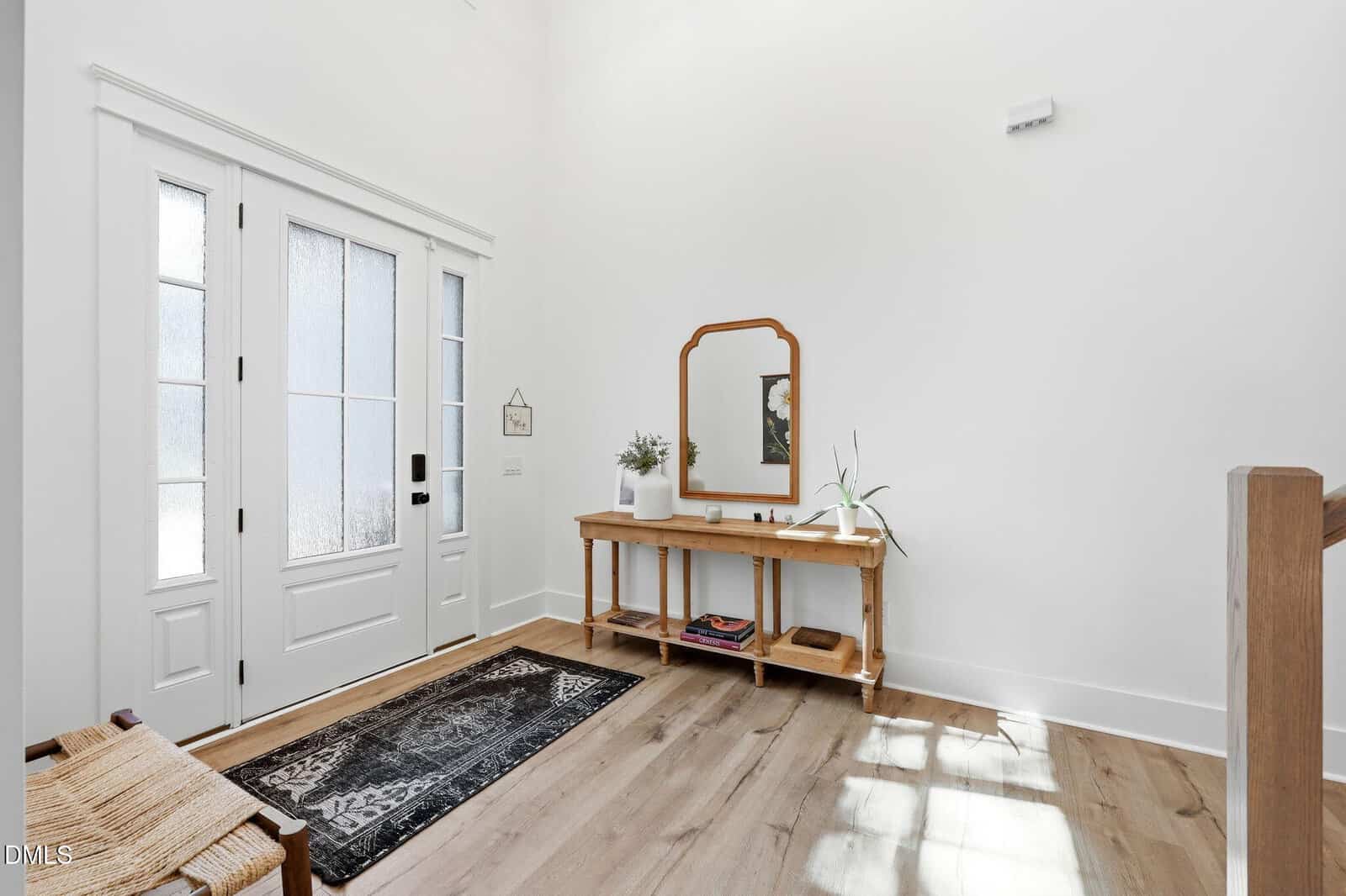 Bright entryway with white walls, wooden floor, a rug, a long console table with a mirror, plants, and decor. Sunlight streams in from glass-paneled front door on the left.