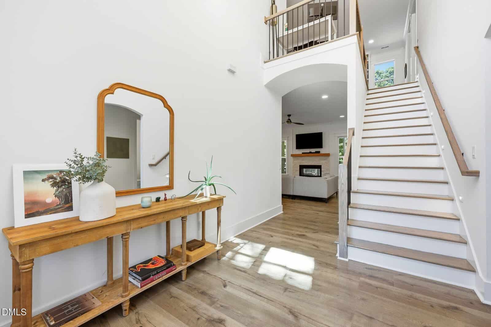 Bright entryway with a wooden console table, decorative items, and a mirror. White walls, hardwood floors, and stairs with a white banister lead to the second floor. Living room with a TV visible in the background.