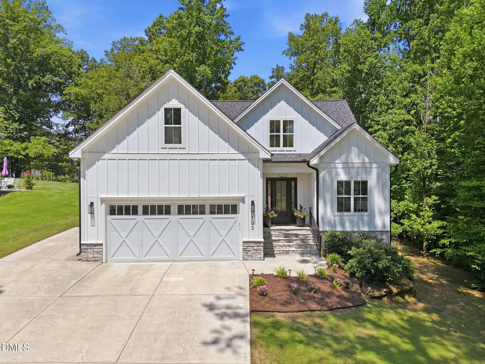 A modern white house with a two-car garage, gabled roof, and neatly landscaped front yard, surrounded by green trees and a wide concrete driveway on a sunny day.
