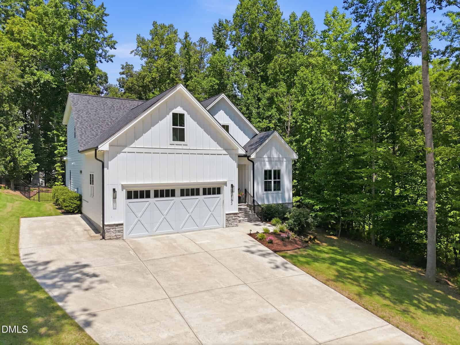 A modern white house at 103 Blue Finch Court, Youngsville, with a two-car garage, gabled roof, and concrete driveway sits surrounded by green trees and a manicured lawn under a clear blue sky.