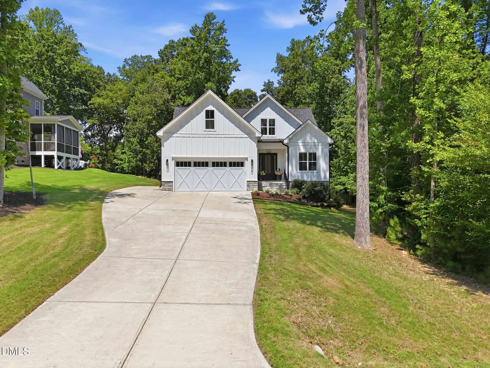 A white modern farmhouse with a double garage sits at the end of a long curved driveway, surrounded by green grass, trees, and neighboring homes under a clear blue sky.