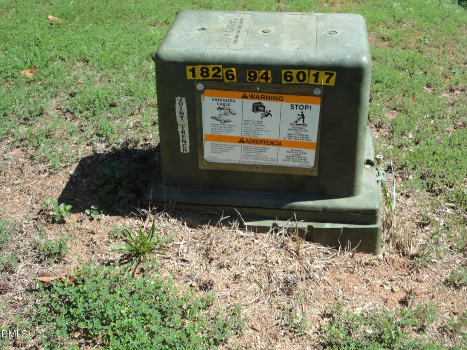 A green utility box with warning labels and the numbers 1826 94 6017 sits on grass and dirt, surrounded by weeds.