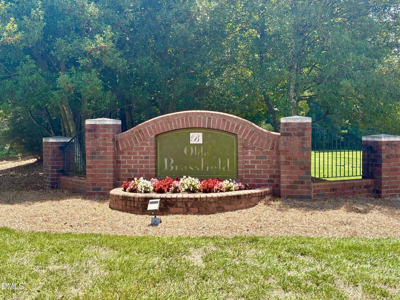 A brick sign with "Olde Brassfield" surrounded by red and white flowers, set against a backdrop of trees and green grass, marks the entrance to 3669 Brooks Bend in charming Franklinton.