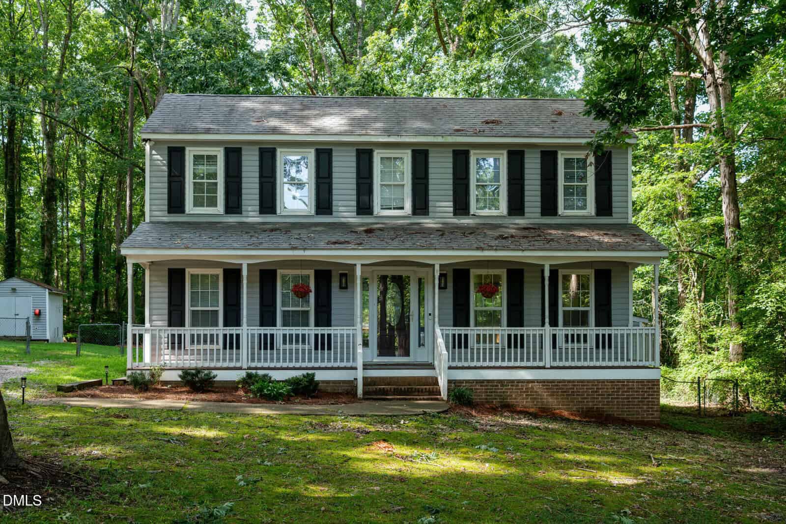 A two-story house at 1011 Homestead Place in Zebulon features gray siding, white trim, black shutters, and a wide front porch with white railings, all surrounded by trees and grass.